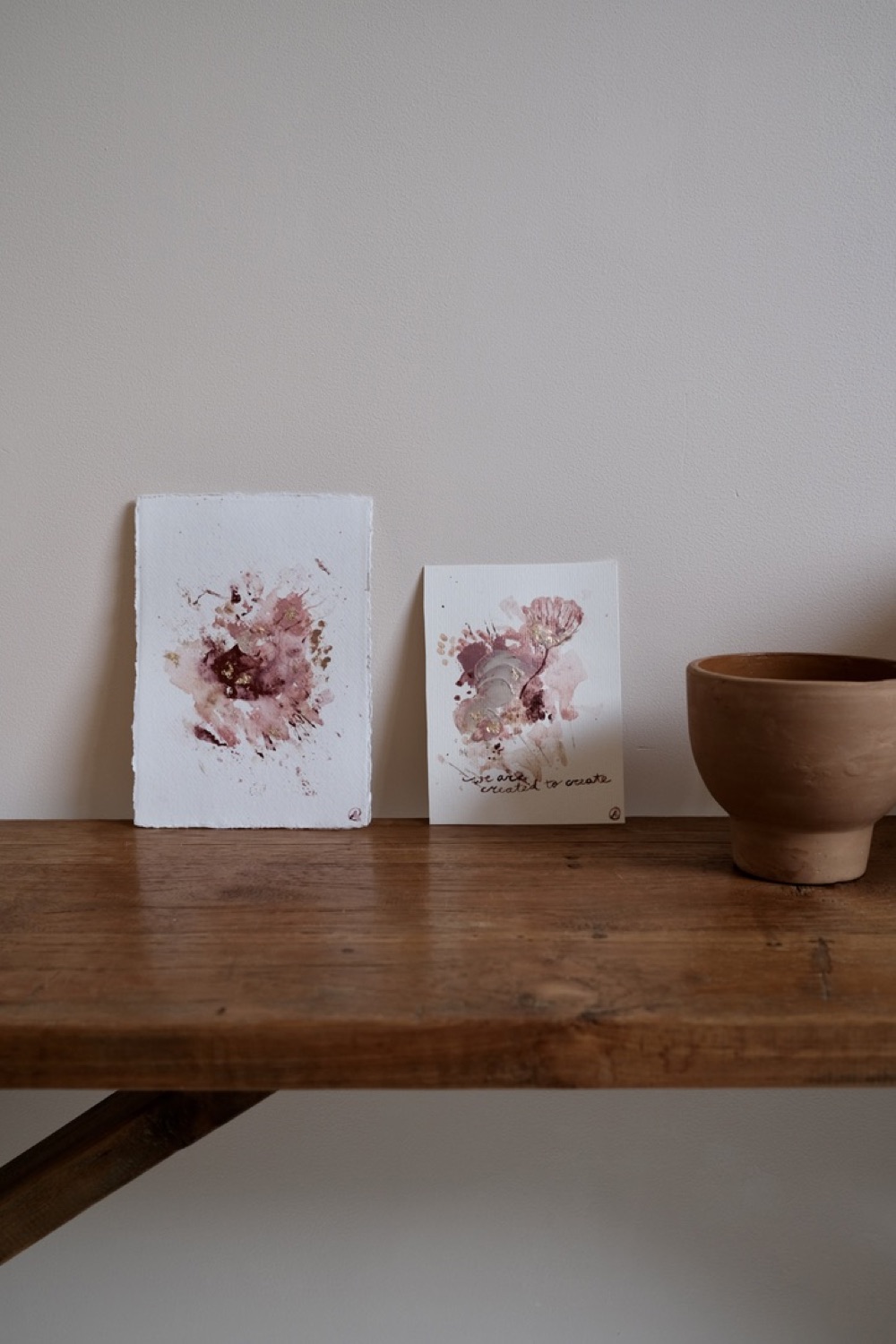 Twee abstracte schilderijen in roze tinten op een houten plank, naast een terracotta pot
